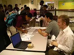 Two young white men sit at a table with several teenage African American students mingle around it, and one signs a paper on it. Also on the table is a laptop.