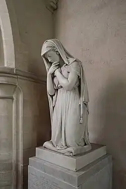 This image shows a white marble monument by Sir Francis Chantrey (1820) commemorating Lady Charlotte Finch at Burley-on-the-Hill, Rutland. The sculpture depicts a standing female figure in classical drapery, her head covered with a veil and bowed in an attitude of profound grief. Her right hand is raised to her face in a gesture of mourning, whilst her left arm hangs at her side. The figure is draped in flowing robes that fall in naturalistic folds to the base, and she stands barefoot upon a simple rectangular plinth. The monument is positioned within a Gothic Revival architectural setting with stone arches visible in the background. This work exemplifies the neoclassical style's emphasis on emotional expression and idealized classical form, demonstrating Chantrey's mastery in conveying deep sentiment through marble. The sculpture's pose and drapery treatment reflect the influence of classical antiquity whilst the overall composition creates a powerful sense of maternal grief and loss.