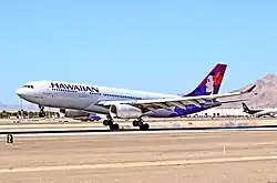 A white twin-engine plane painted with the word "HAWAIIAN" in the front and a woman in different purple hues on the tail has just landed on a runway on a sunny day amid a desert backdrop