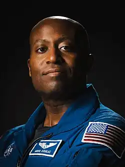 An African American man in a blue flight suit is standing in front of a dark background; he is facing the camera and smiling.