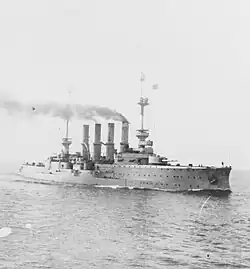 A large ship steams through calm seas, thin black smoke drifts up from its smoke stacks