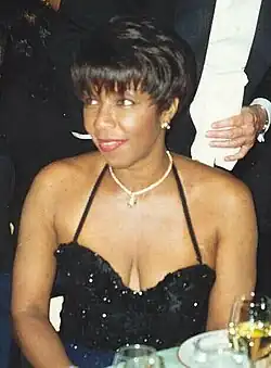 The head and shoulders of a woman, wearing a neckless and a low-cut black dress.