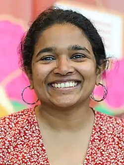 Headshot of Netha Hussain behind a mural. She is a young woman with her hair tied up wearing a red flower dress.