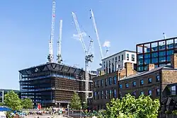 Four white construction cranes over a large brown structure under a blue summer sky. A square with people in the foreground. To the right are four other buildings: Two buildings of steel and glass loom over two brown brick buildings.