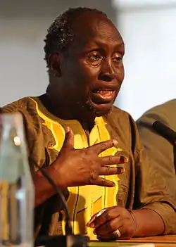 Headshot of Ngũgĩ wa Thiong'o speaking at a literature conference. He, then, was an elderly black man with dark, curly hair and a stubby goatee, wearing a light yellow shirt.