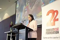 Livingstone speaks at a lectern. She wears a white jacket and is smiling as she speaks. A banner for the Bob Rose Foundation stands next to her and a photo of Australian Rules Football players is visible on a projector screen behind her.