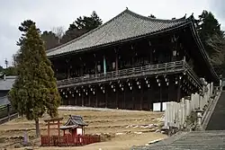 Large wooden building with a pyramid shaped roof. Built on a hill slope with part of the building and veranda supported by poles.