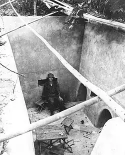 A soldier sitting on a chair in a large exposed underground room. To the right are two large holes in the wall while the room is littered with other furniture, including a makeshift table.