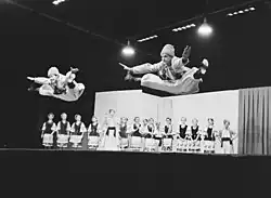 2 male dancers dressed in Ukrainian-style folk clothing jump in the air. A group of female dancers, also in Ukrainian-style folk clothing, watch them from the stage.