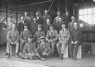 Group portrait of nineteen men, including six in military uniforms with peaked caps, in front of a biplane in a hangar