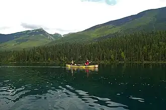 Two paddlers sit in a canoe, gently paddling across the calm water. Behind them, large green mountains flank the water.