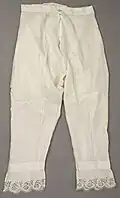 Vintage white pantalettes with an open-crotch design, featuring a button closure at the waist and floral lace trim at the cuffs, showcasing historical practicality and decorative elegance, 1830s