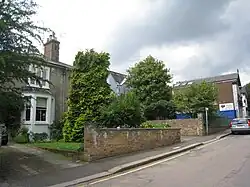 Photograph of several houses, partly hidden by trees, behind a garden wall on an upward-sloping suburban road