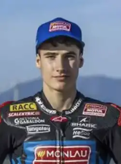 slim young caucasian male at a race track dressed in grey leather racing jacket emblazoned with sponsors' logos and blue baseball cap