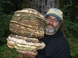 Photo of a bearded and bespectacled white man in the woods; he is holding a fungus that is several times larger than his head