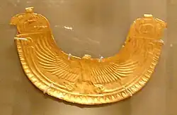 Image of a gold pectoral in the shape of an eagle spreading its wings. The wings end in two Horus heads at the tip of each of the wings
