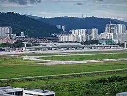 Aerial view of an airport, with factories at the foreground. More buildings and hills form the backdrop.