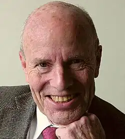 Colour publicity portrait of a smiling, clean-shaven man with receding hair, resting his chin on his hand. He is wearing a tweed jacket, white shirt and red tie.