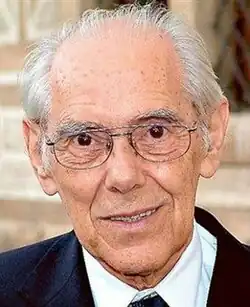 Colour Press shot of an elderly, half-smiling, bespectacled, clean-shaven man, with receding short white hair, brushed back.