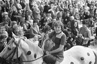 A black and white photograph of a young girl riding a grey horse with black spots painted on it.