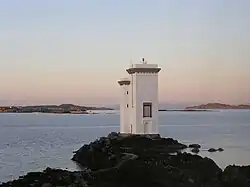 A white building consisting of a single white tower four stories high and an adjacent tower appearing to be a single storey lower, sits on a rock overlooking the sea. The sun is not visible but is evidently low in the sky as pinkish light suffuses the landscape. In the distance there are a myriad low rocky islets.