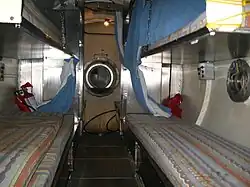 Internal view along the centreline of a cylindrical chamber with four curtained bunk beds in the foreground and a open lock door in the background.