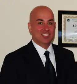 Pedram Salimpour in a dark suit and tie, smiling, standing in front of a framed certificate on the wall.
