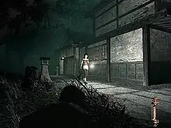 A horizontal rectangular video game screenshot that is a digital representation of the exterior of a traditional Japanese house in a village. A girl uses a flashlight to illuminate the dark surroundings of a path outside a house.