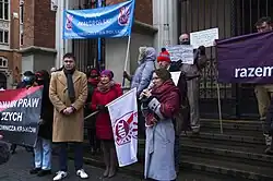 Several protesters holding banners, Gosek-Popiołek speaking to a microphone, "Left Together" flag visible behind her