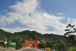Photograph of a bridge leading to a hill. Two construction workers are working on the bridge, with a sign in Spanish that reads "Warning! Men working" before the bridge.