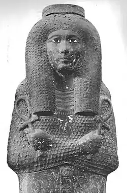 An Egyptian coffin with the carved image of a woman's face and body on the lid. Her arms are crossed, and she is portrayed as holding an ankh in each hand (a key of life symbol, shaped like a "T" with a loop on top).