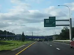 Ground-level view of a freeway; a large, green sign marked with "Exit 7" is posted directly overhead. Two large bridges are visible in the distance.