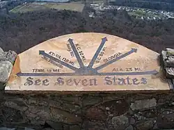 Carved rock plinth inscribed with six arrows and seven state names located high up on the mountain with dark forest beneath and township buildings in distance