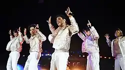 A group photo of SB19 performing, each wearing white clothing; each member has both of their arms raised in the air