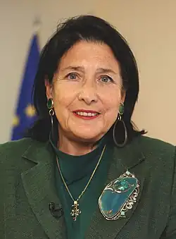 Official photograph of Salome Zourabichvili, a white middle-aged woman facing the camera, wearing a green suit with a cross necklace and standing in front of the European Union flag.