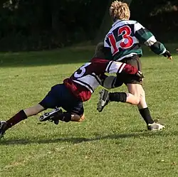 A child running away from camera in green and black hooped rugby jersey is being tackled around the hips and legs by another child in opposition kit.