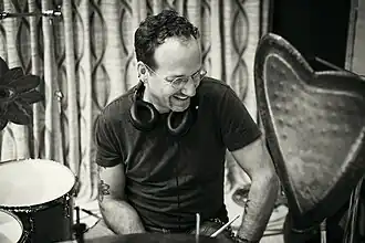 black-and-white image of Scott Amendola wearing a dark t-shirt, glasses, and a set of headphones around his neck, sitting behind a drum kit, looking left of camera and laughing