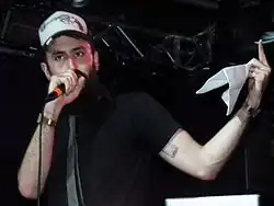 Image of the white, bearded rapper performing on stage, holding a microphone. He is wearing a black t-shirt, a grey tie and a "trucker cap". The cap has a pistol morphed into a microphone.
