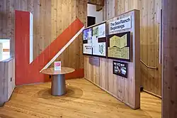 Photograph of visitor information area at Sea Ranch Lodge that includes a graphic display titled "The Sea Ranch Beginnings". The space is finished with wood plank. There's a large red arrow painted on the wall.