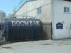 A large, cylindrical tank with the words Doom Bar written on it, lying horizontal behind the fence of an industrial unit