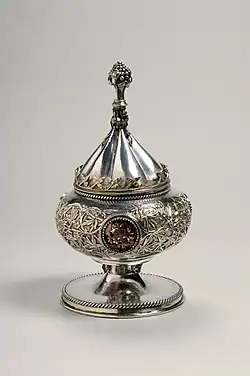 Silver gilt vessel with conical lid. It was intended for use as a ciborium. The vessel is decorated with foliate scrolls and medallions. The knob on the vessel's lid is in the shape of a bunch of grapes.