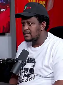 A man seated behind a microphone, wearing a black baseball cap and white t-shirt