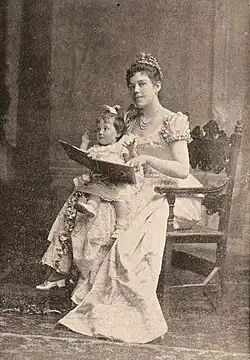 Woman wearing a ruffled gown and tiarra, seated on a chair, and holding a child and a book