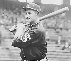A man in a dark baseball uniform and cap poses with a baseball bat on his shoulder.