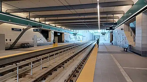 The below ground Spring District station on opening weekend in April 2024. The station has a high ceiling with exposed beams and bright lights. The station has two platforms on the sides of the station with two tracks running down the center. There are benches along the platform for passengers to wait. The walls are decorated with artwork, and there are signs with information about the station and train schedules.