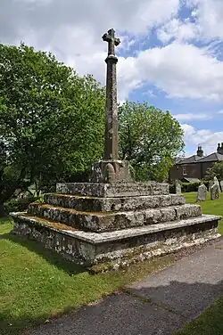 A stone cross on a chamfered column, on four stone square steps, with trees in the background