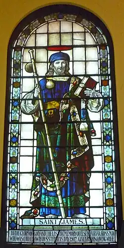 St James is shown in layered robes holding a staff across his body and a Bible in his left hand. On his right shoulder is the scallop shell.