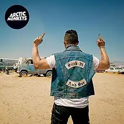 A shot of drummer Matt Helders from behind wearing a denim jacket with the title of the song on it, while holding up two middle fingers. The band name is on a black circle in the top left corner.