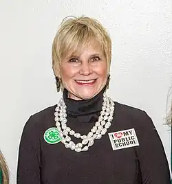 A white woman with blonde hair is wearing a brown turtleneck and a large white necklace; she is looking and smiling into the camera.