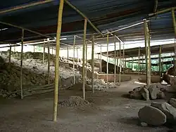 A cleared area of bare soil strewn with rubble, with raised areas to the left and right and roofed with corrugated metal supported by timber poles